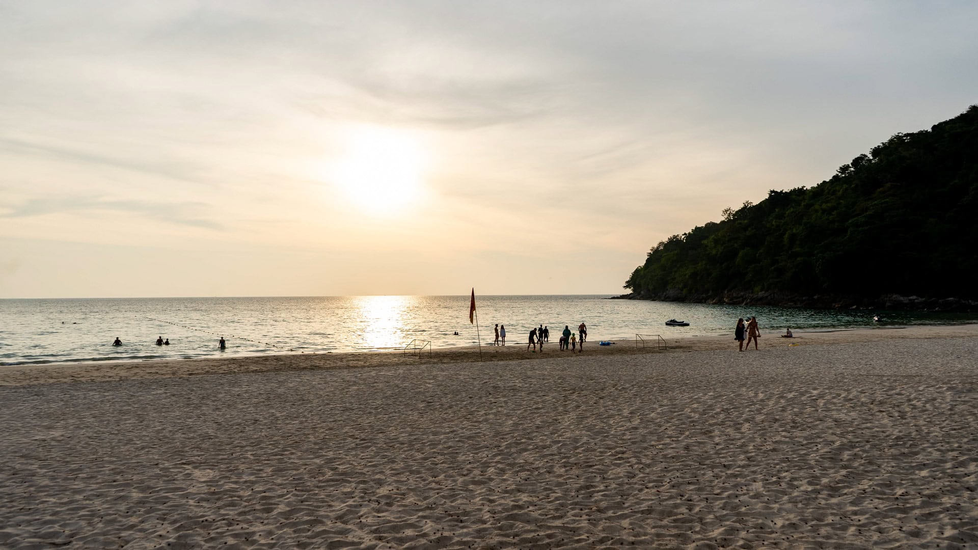 Karon Noi Beach Phuket at sunset with calm sea, people swimming near shore, and low sun over the horizon