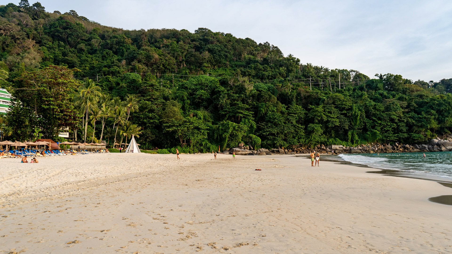 Karon Noi Beach Phuket showing wide sandy shoreline, scattered visitors, and dense green hillside at the back of the beach