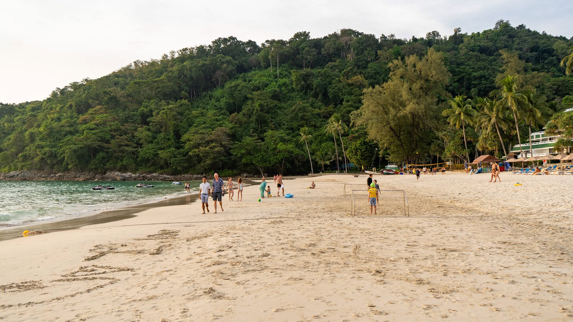 Karon Noi Beach Phuket with people walking along the waterline, small boats offshore, and trees lining the beach