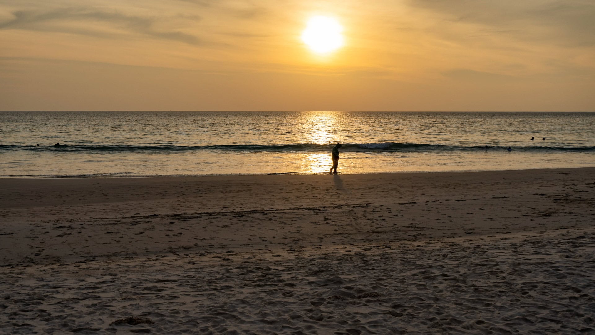 Karon Noi Beach Phuket at sunset with a person walking along the shoreline and sunlight reflecting on the sea